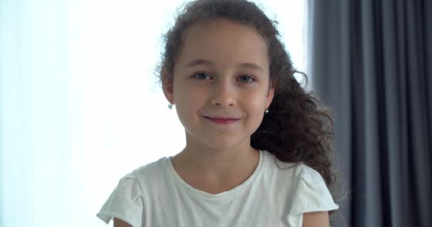 Porträt Lustiges kleines Mädchen lächelndes Kind, das in die Kamera schaut, sitzt zu Hause auf der Couch, süßes Kind mit hübschem Gesicht sitzt auf dem Sofa. Konzept einer glücklichen Kindheit. — Stockvideo