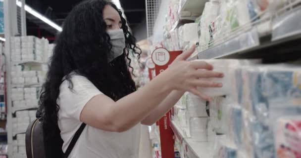 Jonge vrouw met een masker van een coronavirus epidemie koopt toiletpapier in een supermarkt, waar mensen paniekerig alles kopen. Corontin, isolatie van mensen. — Stockvideo