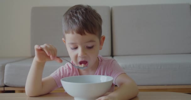 귀여운 사내 아이가 집 소파에 앉아 있는 물 한 컵을 마시고 있습니다. 작은 귀여운 아기가 식탁에 앉아 직접 오트밀을 먹고 있는 동안, 아기는 기꺼이 먹는다. 개념 상의 행복 한 어린 시절. — 비디오