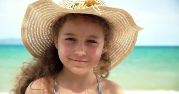 一个戴着帽子的滑稽小女孩的画像，一个看着摄像机的微笑的孩子，站在海边，一个长着漂亮脸蛋的可爱孩子站在海滩上。快乐童年的概念. — 图库视频影像