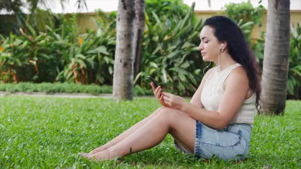 Mujer uso por teléfono móvil escuchar música en la naturaleza relajarse en el parque sentado en el césped o césped en la calle. — Vídeos de Stock