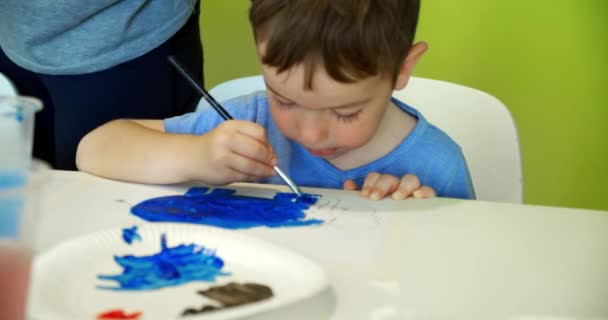 Ευτυχισμένα χαριτωμένα παιδιά, ένα κορίτσι 7-8 ετών και προσχολικής ηλικίας αγόρι, χρώμα στο σπίτι με ακουαρέλες κάθεται σε ένα λευκό τραπέζι, μικρό αγόρι ζωγραφίζει σε χαρτί με έγχρωμες μπογιές και το κοριτσάκι στο λευκό σορτς της — Αρχείο Βίντεο