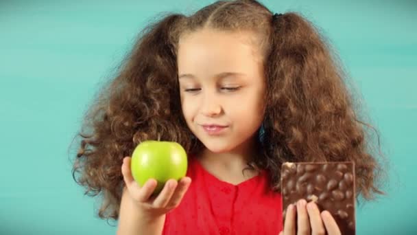 ターコイズのかわいい子供グリーンアップルとチョコレートを保持し、果物とチョコレートの間で健康的な食事を選択する背景。 — ストック動画