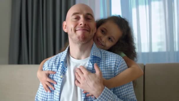 Día del Padre.Acercamiento retrato de un padre y su hija que se sientan junto a un abrazo mirar a la cámara y el uno al otro en los ojos sentados en el sofá en casa.Padre sostiene a su hija a mano abrazos — Vídeos de Stock