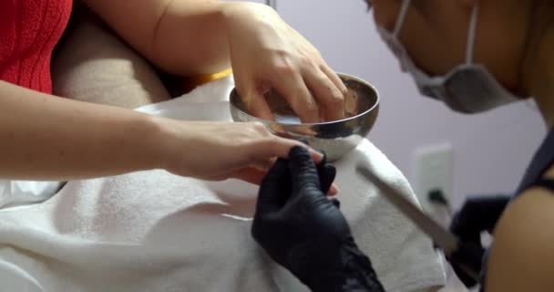 Kosmitolog dělá manikúru brunetce v kadeřnickém salónu. Žena kosmetička provádí zákrok, připravuje a zpracovává klienty nehty se speciálním skalpelem k odstranění starého laku na nehty. — Stock video