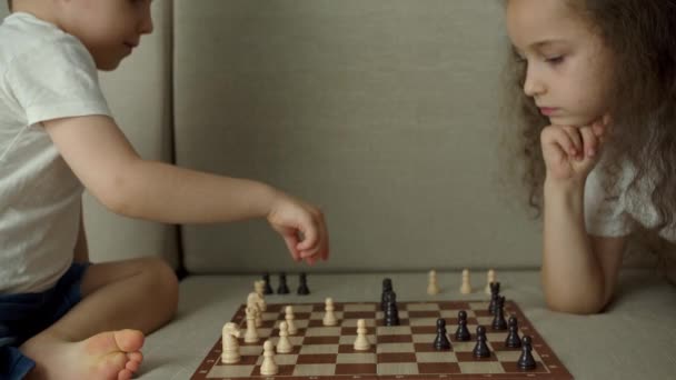 Desenvolvimento do pensamento lógico. Retrato de um rapazinho esperto e irmã mais velha a jogar xadrez. As crianças jogam xadrez enquanto estão sentadas no devan em casa, o desenvolvimento do pensamento lógico nas crianças. — Vídeo de Stock