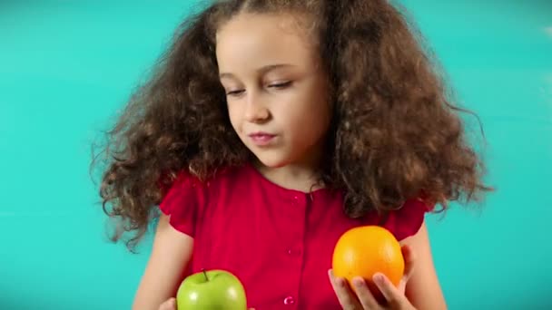 Happy Kid gör ett val till förmån för ett grönt äpple. Roligt barn på turkos bakgrund att hålla ett grönt äpple och apelsin och val hälsosam mat mellan frukt och apelsin. — Stockvideo