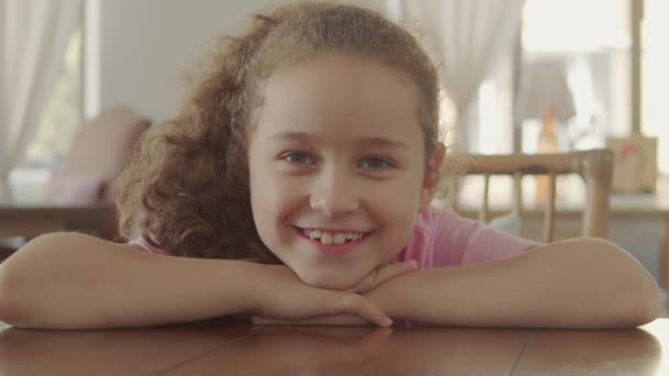 Portret van een gelukkig klein meisje, lachend kind, kijkend naar de camera, zittend aan tafel in een café, schattig baby kind met een mooi gezicht, kind zittend aan tafel thuis. Gelukkige jeugd concept. — Stockvideo