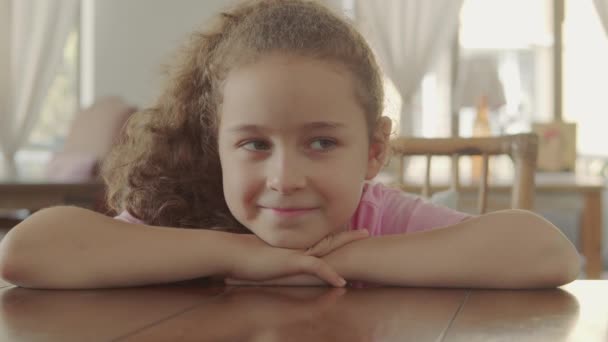 Retrato de una niña feliz, un niño sonriente, mirando a la cámara, sentado en la mesa en un café, un bebé lindo con una cara hermosa, un niño sentado en la mesa en casa. Concepto de infancia feliz. — Vídeos de Stock