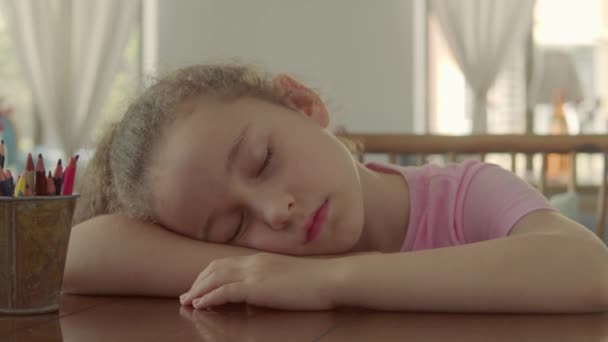 Zblízka tvář malé holčičky, dítě unavené v horkém dni se snaží usnout na boku v náručí, zatímco sedí u stolu. 8-letá dívka u stolu po lekci kreslení. — Stock video