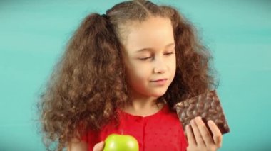 Child Makes a Choice Between of Holding a Green Apple and Chocolate Cute Child on Turquoise Background of Holding a Green Apple and Chocolate and Choice Healthy Eating between Fruit and Chocolate.