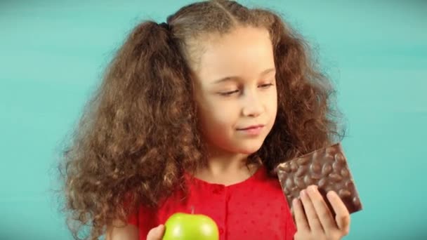 Child Makes a Choice Between of Holding a Green Apple and Chocolate Cute Child on Turquoise Background of Holding a Green Apple and Chocolate and Choice Healthy Eating between Fruit and Chocolate. — Stockvideo