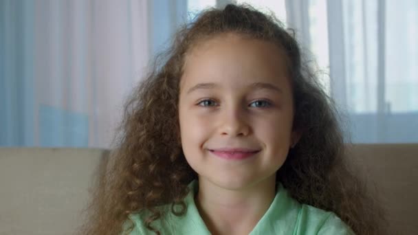 Портрет Медова маленька дівчинка усміхнена дитина, дивлячись на камеру, сидить на дивані вдома, мила дитина з красивим обличчям, сидячи на дивані. Концепція щасливого дитинства . — стокове відео