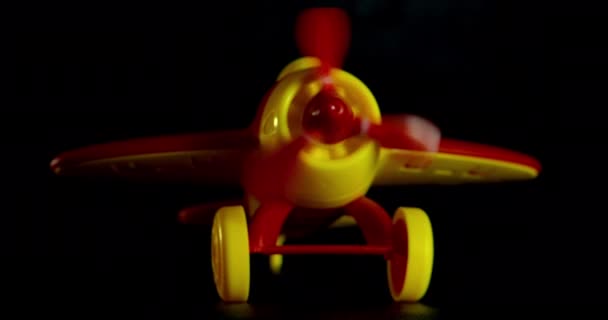 Filmé dans une touche sombre. Plan macro d'un jouet pour enfants avion rouge avec des ailes jaunes et des roues sur un fond noir, l'avion démarre, son hélice tourne et commence à avancer. — Video