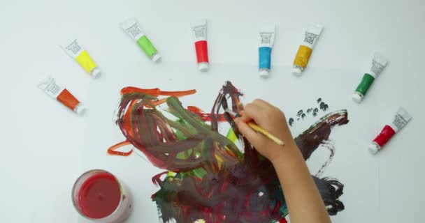 Close-up beeldmateriaal bovenaanzicht van hoe een kleine jongen tekent met een kwast op een wit vel van verschillende kleuren met verf, abstracte tekeningen maakt met een artistieke kwast met behulp van aquarellen. — Stockvideo
