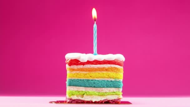 Slow motion, 4K Stock Footage. A slice of birthday cake with a bright blue candle on a pastel pink background. Slow motion, a festive candle is burning on a rainbow piece of cake. — Stock Video