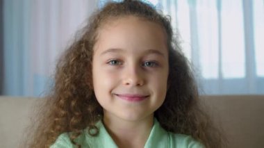 Tatlım, gülümseyen küçük kız evdeki kanepede oturmuş kameraya bakıyor, güzel yüzlü tatlı çocuk kanepede oturuyor. Mutlu bir çocukluk kavramı.