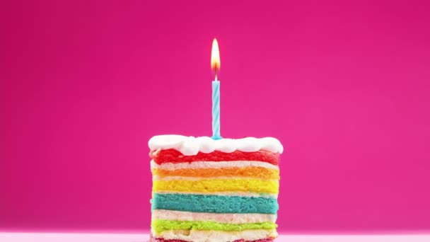 Задувается кусочек праздничного торта с ярко-желтой горящей свечой на розовом фоне. Праздничная свеча горит на радужном куске торта, который угасает и медленно тлеет. — стоковое видео