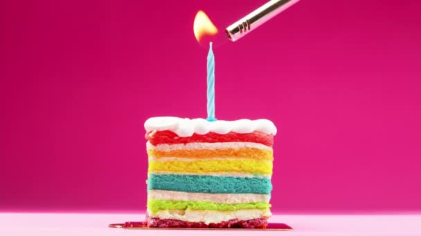 Una rebanada de pastel de cumpleaños con una vela azul brillante sobre un fondo rosa pastel. cámara lenta, una mano de mujer enciende una vela festiva con un pedernal, fotage de la acción. — Vídeos de Stock