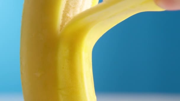 Macro lente tiro banana fresca fica descascando em um fundo azul. Uma mão feminina remove ou remove lentamente a pele de uma banana, conceito de fruta, descascando uma banana. — Vídeo de Stock