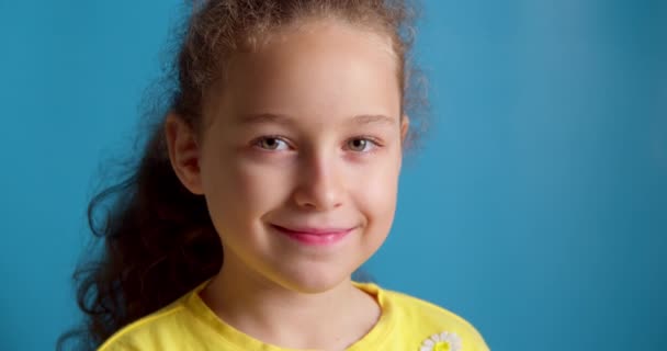 Portrét Legrační holčička usmívající se dítě dívající se do kamery sedí na modrém pozadí, roztomilé dítě s pěknou tváří. Koncept šťastného dětství. — Stock video