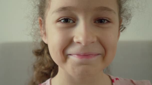 Portrait Drôle petite fille souriante enfant regardant la caméra sont assis sur le canapé à la maison, enfant mignon enfant avec joli visage assis sur le canapé. Concept d'une enfance heureuse. — Video