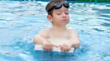 Gözlüklü neşeli çocuk havuzda banyo yapıyor. Yüzme gözlüklü bir anaokulu çocuğu havuza su sıçratıyor, bir çocuk atlıyor ve elinde bir yüzme tahtası tutuyor..