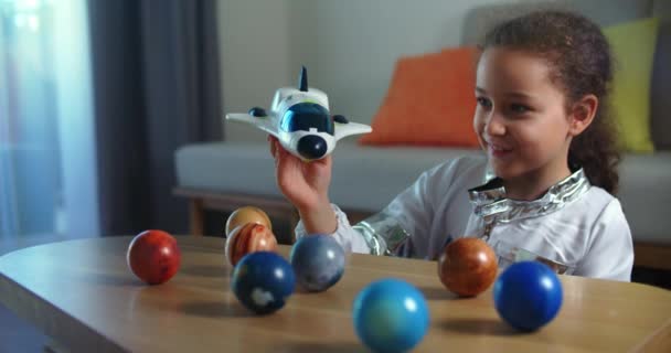Dziecko bawi się w domu astronautą, dziewczynką 8-9 lat w zabawkowym skafandrze kosmicznym, uśmiechnięte dziecko wystrzeliwuje rakietę kosmiczną na tle planet systemu kosmicznego, pilot leci w kosmos — Wideo stockowe