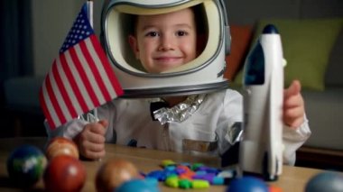 Astronotun evinde oynayan bir çocuk, astronot kostümü giymiş 5 yaşındaki bir çocuğun portresi, kameraya bakan mutlu bir çocuk, yakın plan, Amerikan bayrağı tutan bir pilot, uzaya seyahat eden