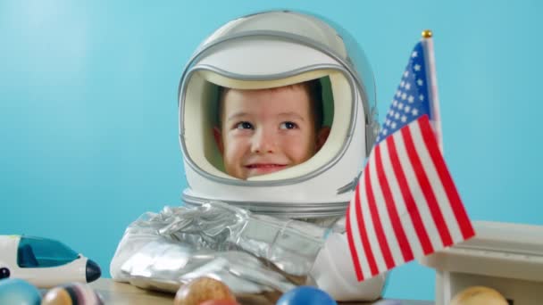 Un niño juega en casa en un astronauta, un retrato divertido de un niño pequeño de 5-6 años en un traje espacial de juguete, un niño sonriente agita la mano, mira a la cámara, de cerca, el piloto vuela al espacio — Vídeo de stock