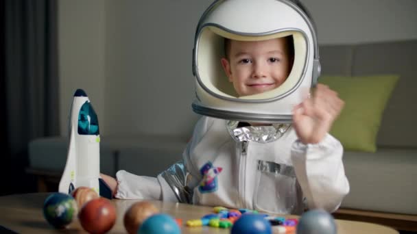 Un niño juega en casa en un astronauta, un retrato divertido de un niño pequeño de 5-6 años en un traje espacial de juguete, un niño sonriente agita la mano, mira a la cámara, de cerca, el piloto vuela al espacio — Vídeo de stock