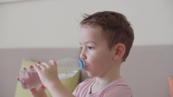 かわいい少年は自宅でソファに座ってボトルから水を飲む。スローモーションの少年飲料水。接近中だ。面白い小さな子供の肖像画水を飲んで. — ストック動画