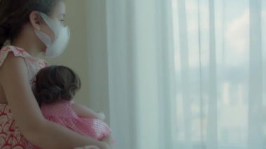 Çocuk kendine ve bebeğine koruyucu maskeler takıyor, yatakta oturup pencereden dışarı bakıyor. Ev karantinasında sıkılmış çocuk. Alerji, covid-19 koronavirüs salgını, salgını önleme.