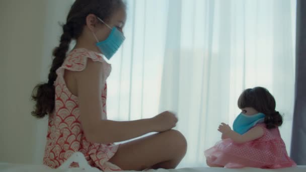 Znudzone dziecko w domowej kwarantannie. Dziewczyna z lalką w ochronnych maskach medycznych siedzi na łóżku i patrzy przez okno. Alergia, pandemia covid-19, zapobieganie epidemii. — Wideo stockowe