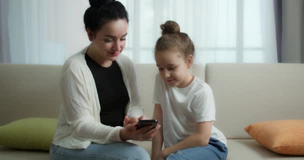행복 한 엄마와 귀여운 딸, 집에서 쉬고 있는 아이와 놀고 있는 엄마는 스마트폰을 사용하여 소파 딸을 껴안고, 재밌는 비디오를 보고, 즐겁게 가족 생활을 즐긴다 — 비디오