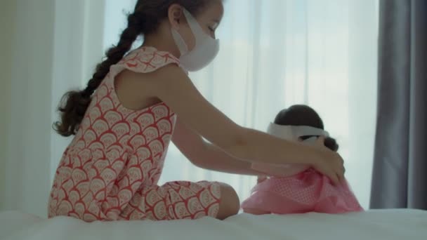 Znudzone dziecko w domowej kwarantannie. Dziewczyna z lalką w ochronnych maskach medycznych siedzi na łóżku i patrzy przez okno. Alergia, pandemia covid-19, zapobieganie epidemii. — Wideo stockowe
