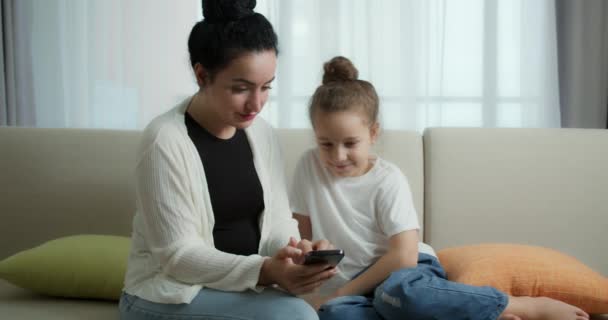 Feliz mamá y linda hija pequeña, madre jugando con el niño en casa relajante usar un teléfono inteligente abrazo sentarse en el sofá hija, ver videos divertidos, divertirse, disfrutar de momentos de la vida familiar — Vídeo de stock