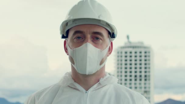 Retrato de un ingeniero profesional de una empresa industrial de construcción que lleva una máscara protectora y traje protector el telón de fondo de grandes edificios altos en construcción. 4k — Vídeos de Stock