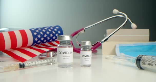 Разработаны вакцина против COVID-19, ампула с вакциной против коронавируса и различных шприцев поблизости, медицинские инструменты, защитная маска, фон американского флага и книги — стоковое видео