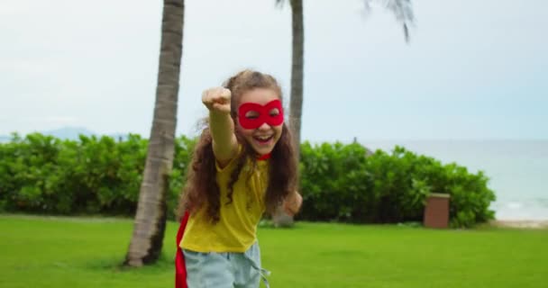 Retrato de un lindo niño divertido, una niña con un disfraz de superhéroe, con una capa roja y una máscara roja, interpreta a un superhéroe, corre por la calle en el parque, demuestra su héroe de fuerza. — Vídeo de stock