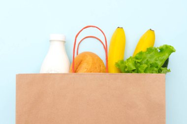 Yiyecek dolu kağıt torba: yeşil salata, ekmek, bir şişe süt, salatalık, muz. Mavi arka plan, üst görünüm üzerine uzan