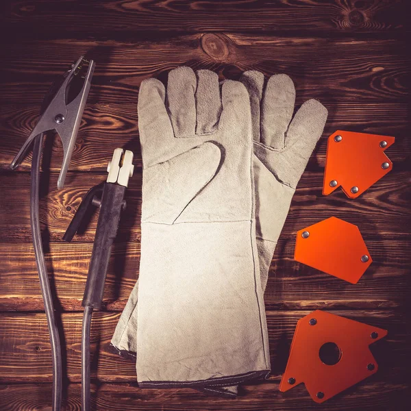 Welding wires, electrode holder and earth terminal, white leather welding gloves, magnetic welding corners on a wooden background. Studio photo in hard light.