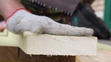 Bir adam paslı demir testeresiyle tahtayı kesiyor ve eldivenli bir elle talaş temizliyor.