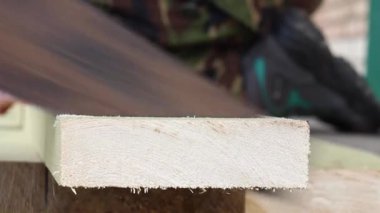 Bir adam paslı demir testeresiyle tahtanın bir parçasını keser ve eldivenli eliyle talaş toplar.