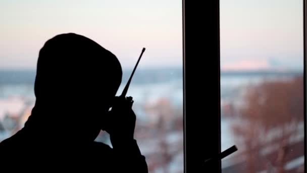 silhouette of a man in a hood stands in front of the window talking into a walkie-talkie