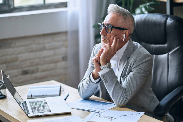 Thoughtful businessman working on laptop while sitting at workplace and using wireless headphones. Business career concept. Copy space