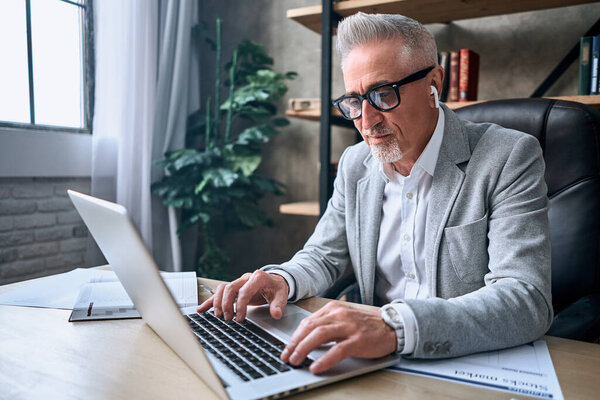 Smiling adult man in glasses working in the office while typing on laptop while using wireless headphones. Business career concept