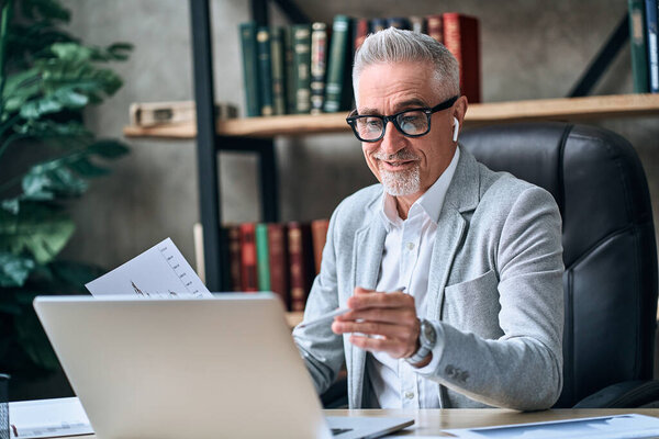 Smiling adult businessman communicating with colleagues during an online call in the office while using wireless headphones. Business concept