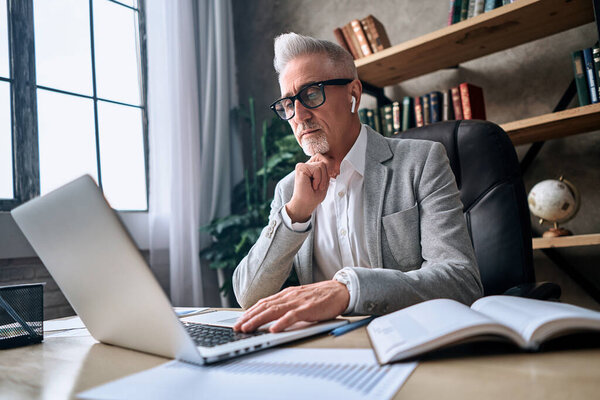 Mature serious man in glasses working online on computer while sitting in his office and holding a hand near the chin. Business concept