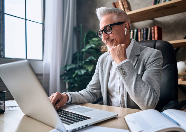 Happy adult businessman using wireless headphones while typing on laptop and looking at screen at hia workplace. Business concept